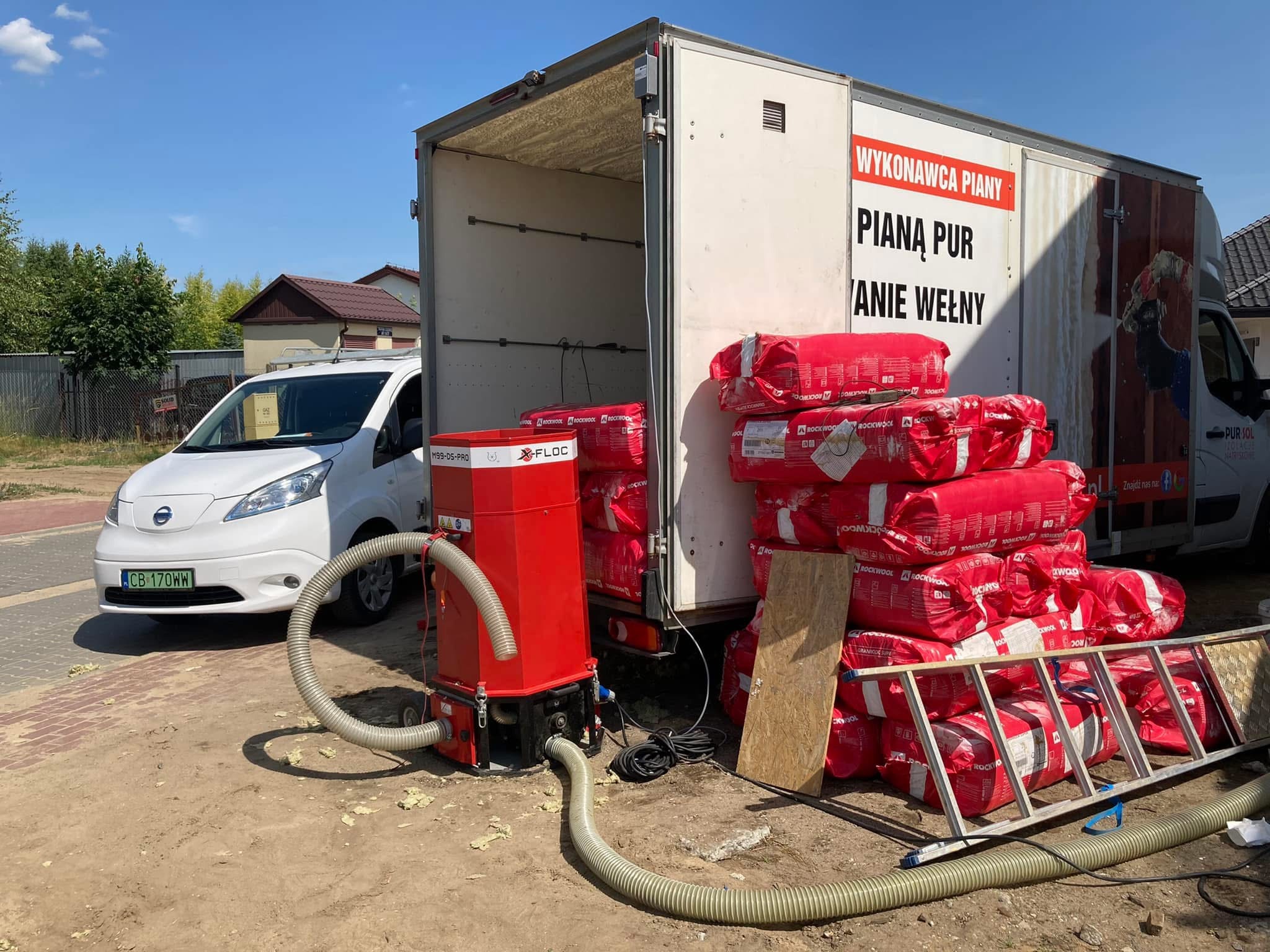 Czerwony agregat do piany PUR obok otwartej furgonetki z workami izolacji Rockwool. Na boku furgonetki napis 'Wykonawca piany PUR'.