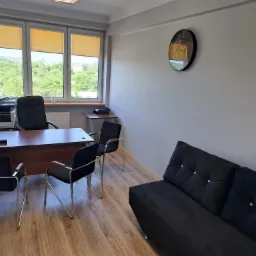 Biuro z drewnianym biurkiem, czarnymi krzesłami z chromowanymi nogami, czarną sofą z guzikami i widokiem na zieleń za oknem.