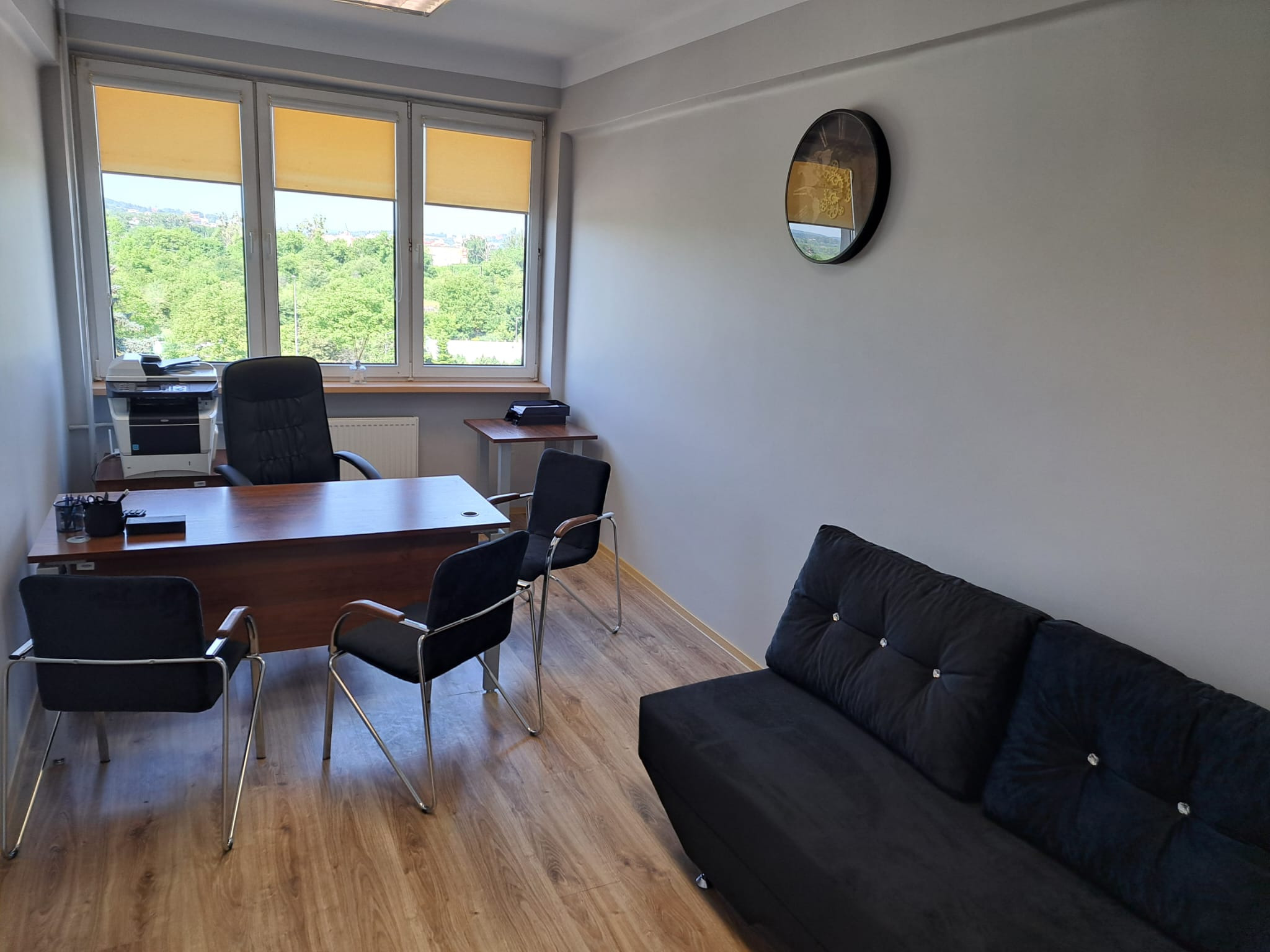 Biuro z drewnianym biurkiem, czarnymi krzesłami z chromowanymi nogami, czarną sofą z guzikami i widokiem na zieleń za oknem.