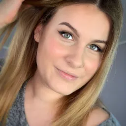 Uśmiechnięta blondynka z delikatnym makijażem oczu i podkreślonymi kośćmi policzkowymi, dotyka włosów dłonią z jasnoróżowym manicure.