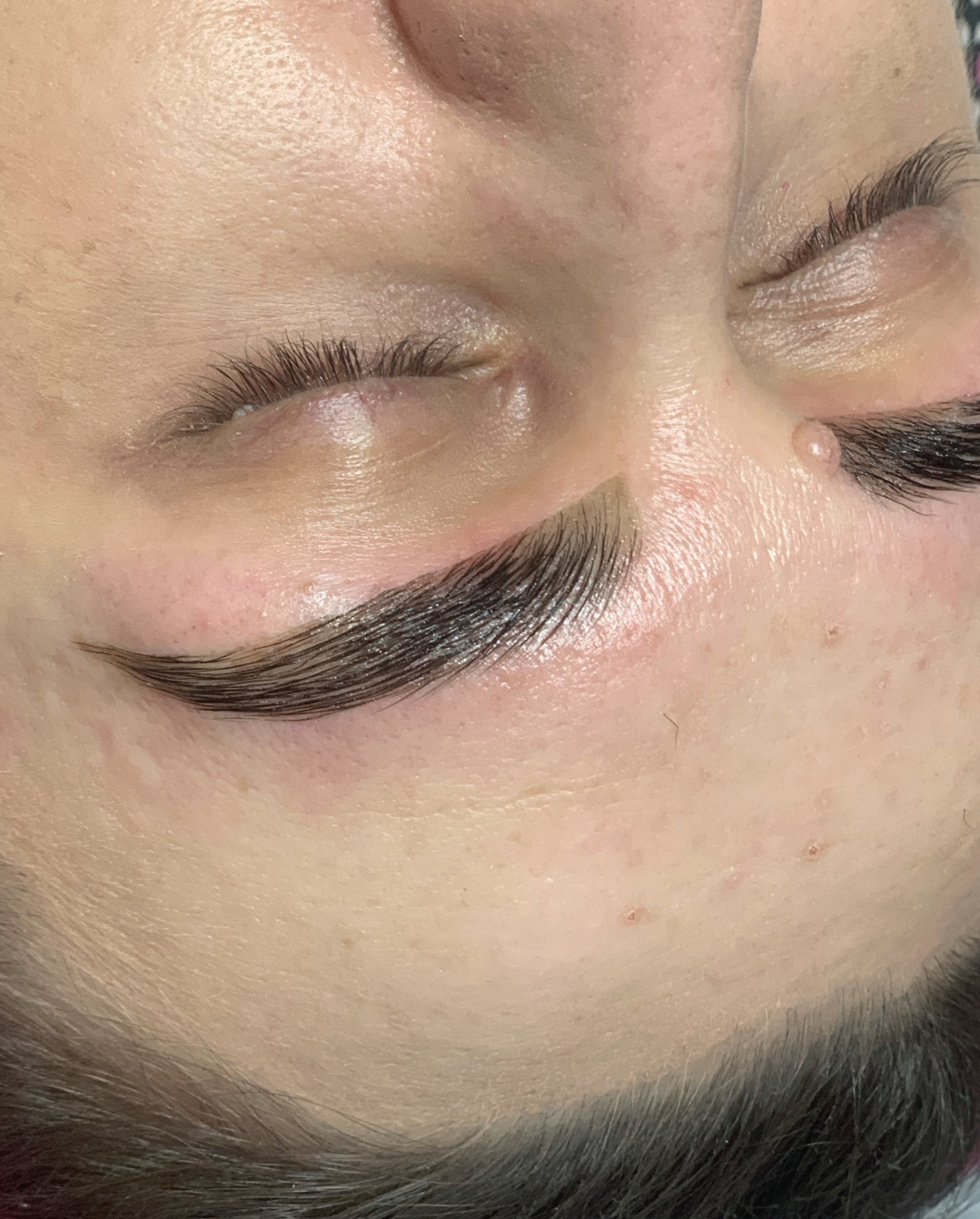 Zbliżenie na brwi po laminacji i koloryzacji, widoczne pojedyncze włoski, skóra z niedoskonałościami, ciemne włosy u nasady.