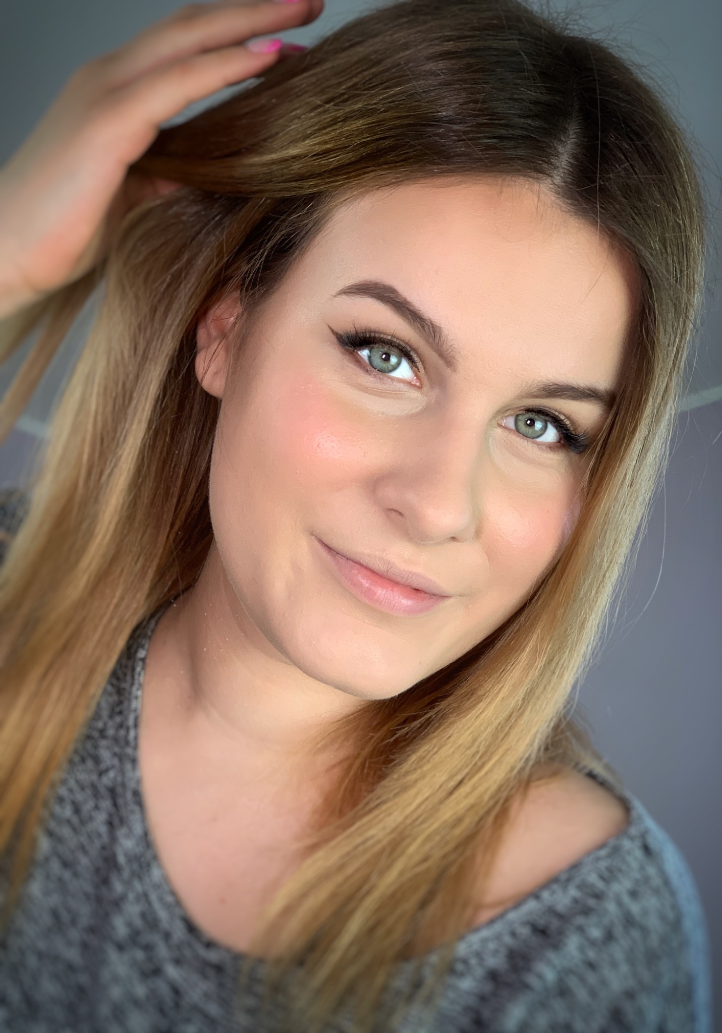 Uśmiechnięta blondynka z delikatnym makijażem oczu i podkreślonymi kośćmi policzkowymi, dotyka włosów dłonią z jasnoróżowym manicure.