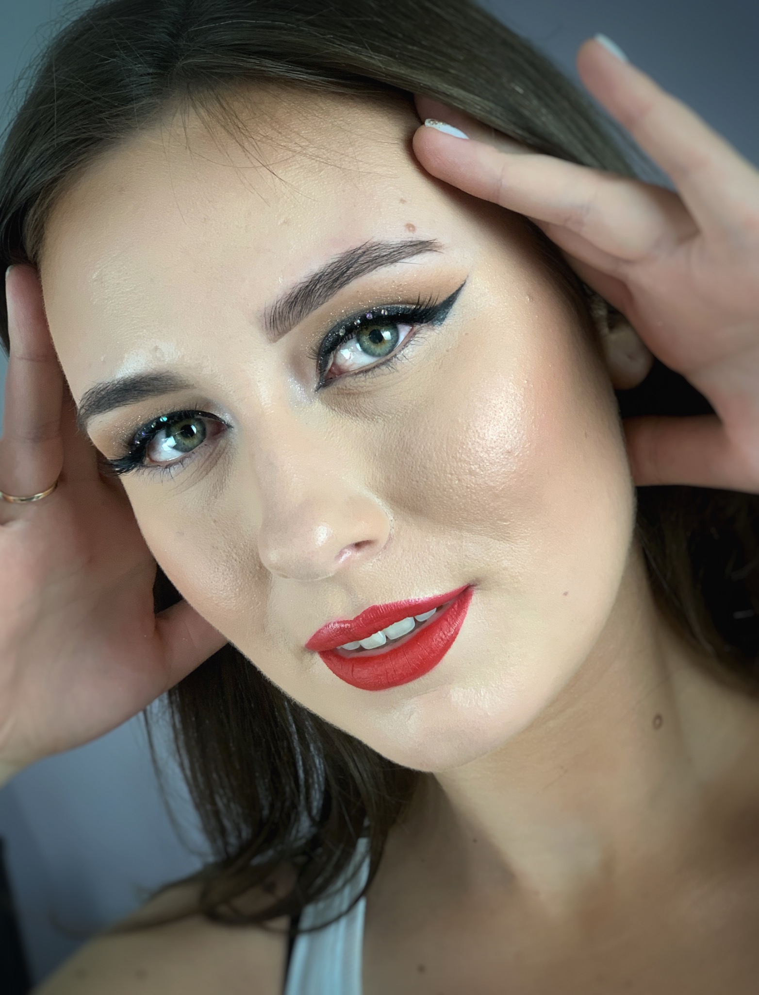 Portret kobiety z intensywnym makijażem: czarna kreska na powiece z brokatem, czerwone usta, brwi podkreślone, dłonie dotykają skroni