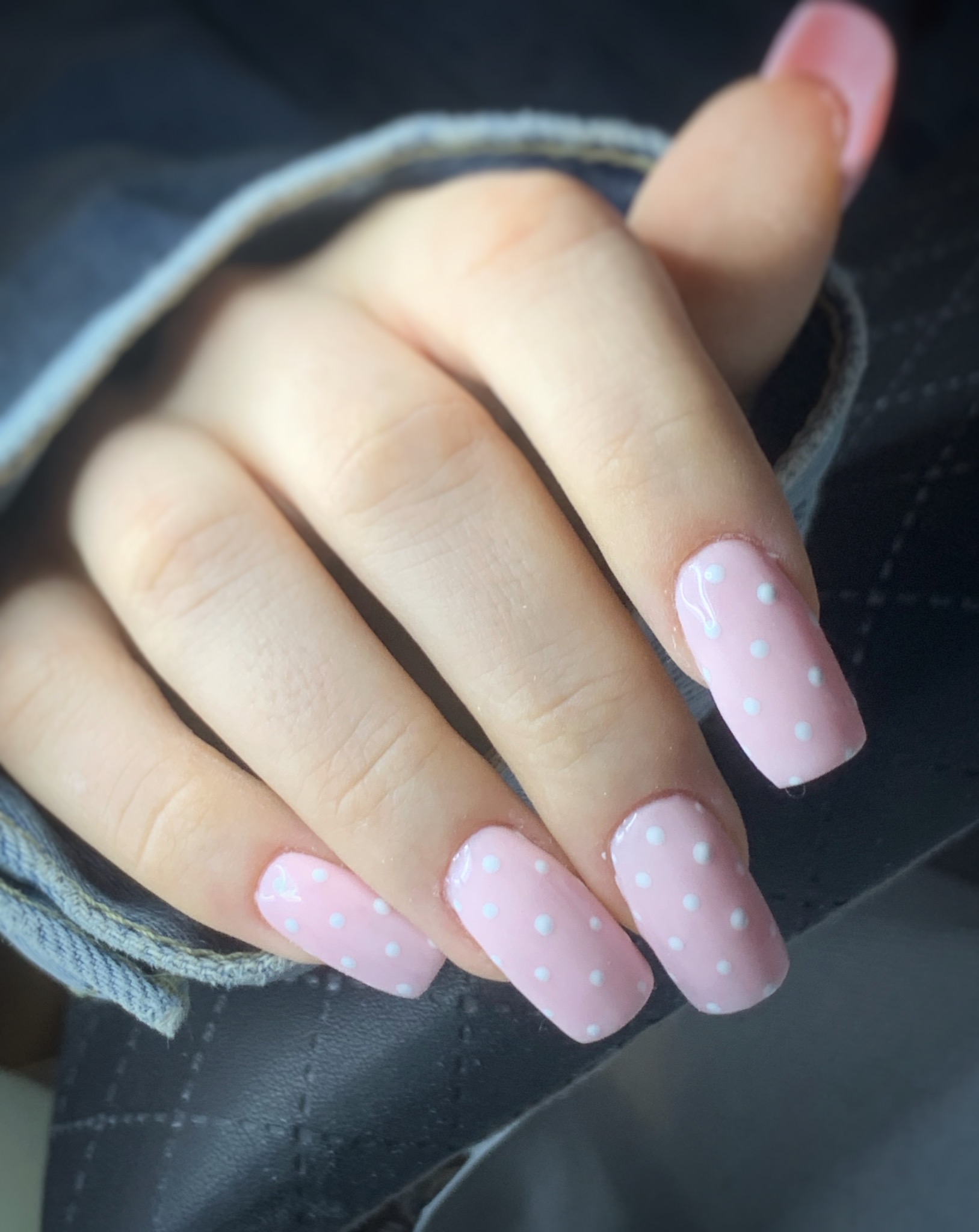 Dłoń z przedłużonymi, kwadratowymi paznokciami pomalowanymi na jasny, pudrowy róż z drobnymi białymi kropkami, widoczny mankiet dżinsowej kurtki w tle.