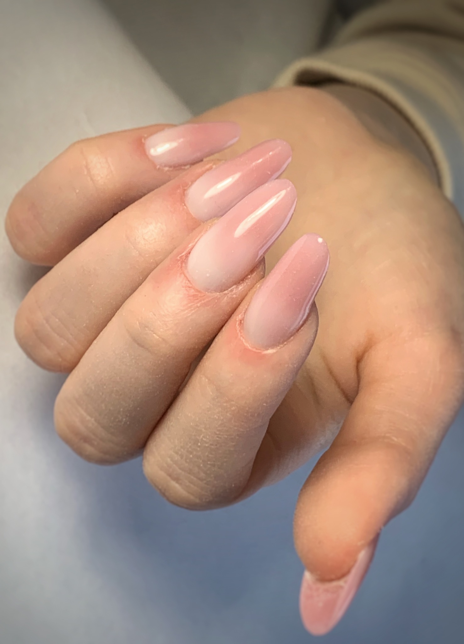 Dłoń z delikatnymi, różowymi paznokciami o owalnym kształcie, z efektem ombre przechodzącym od bieli u nasady do jaśniejszego różu na końcach.