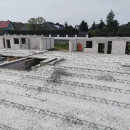 Budowa domu jednorodzinnego w trakcie wylewania stropu, widoczny pracownik w pomarańczowej kamizelce na tle zabudowy mieszkalnej.