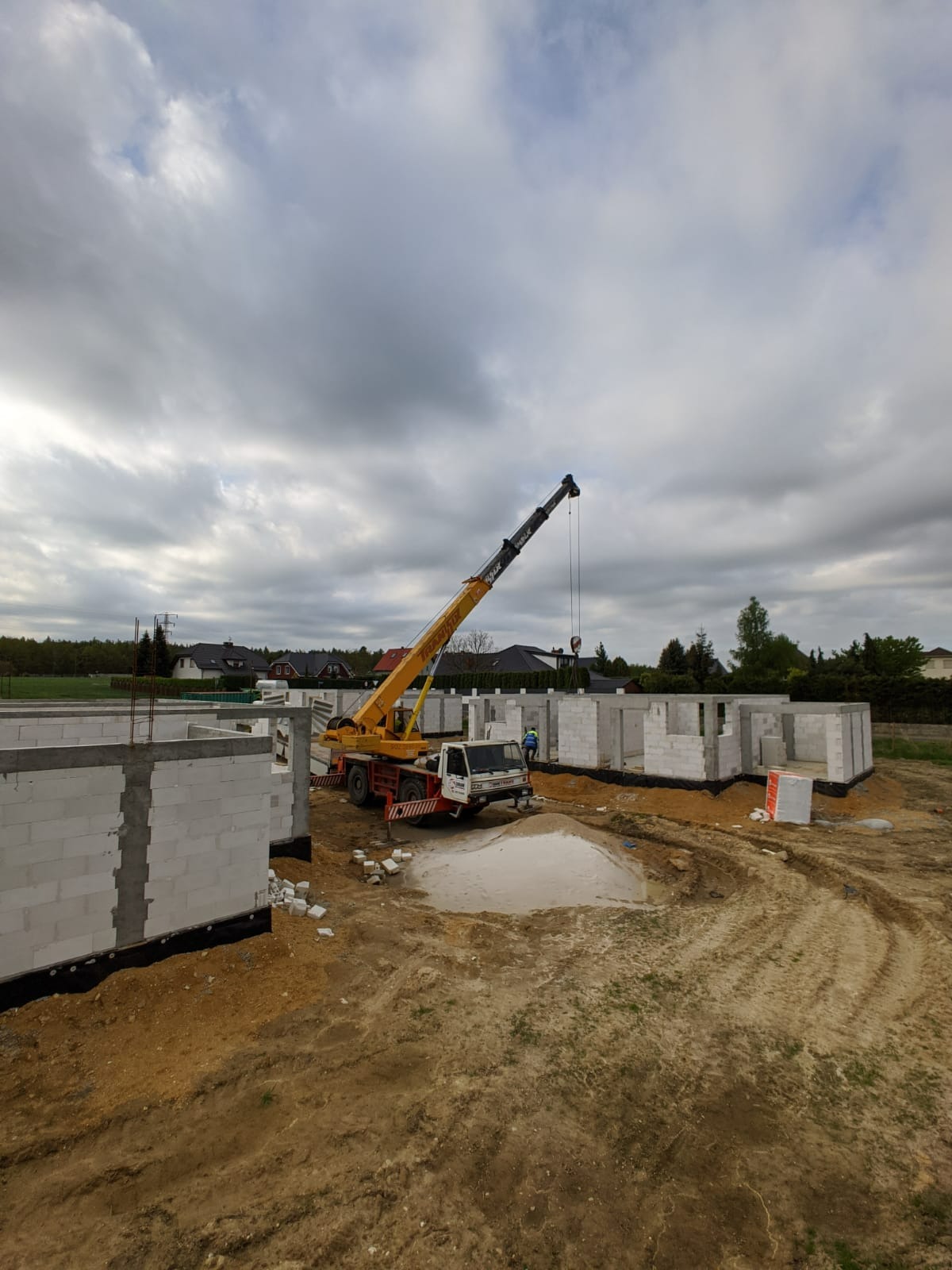 Wznoszenie ścian parterowego domu z bloczków betonu komórkowego w Głogowie, widoczny żuraw budowlany i robotnik w oddali, pochmurne niebo.