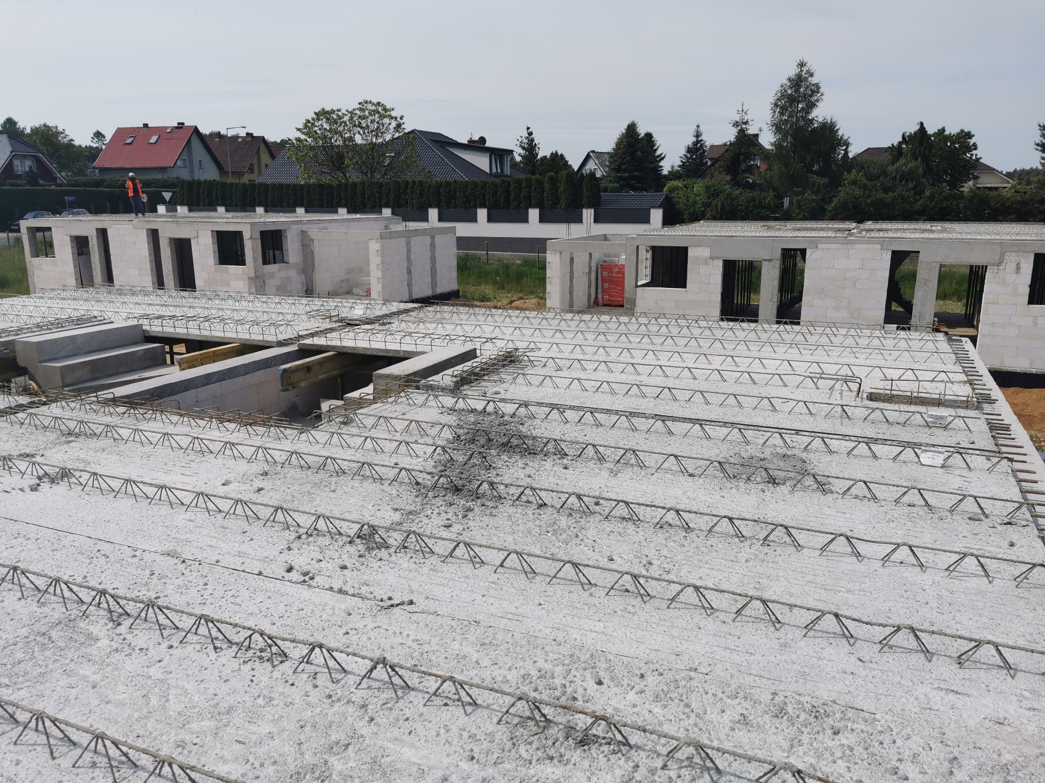 Budowa domu jednorodzinnego w trakcie wylewania stropu, widoczny pracownik w pomarańczowej kamizelce na tle zabudowy mieszkalnej.