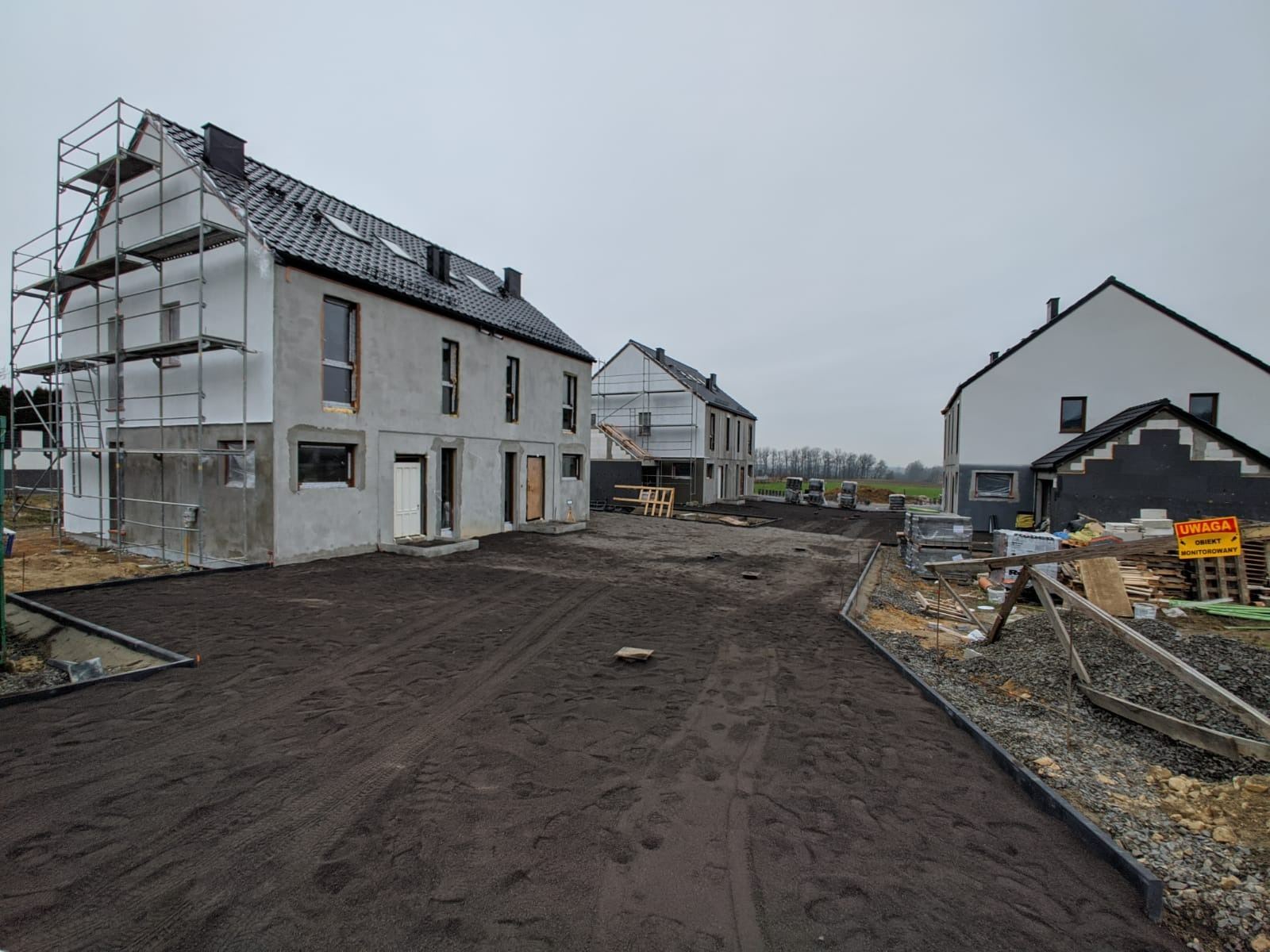 Nowe domy w zabudowie bliźniaczej w trakcie budowy, z widocznymi rusztowaniami, czarną nawierzchnią podjazdu i tablicą ostrzegawczą.