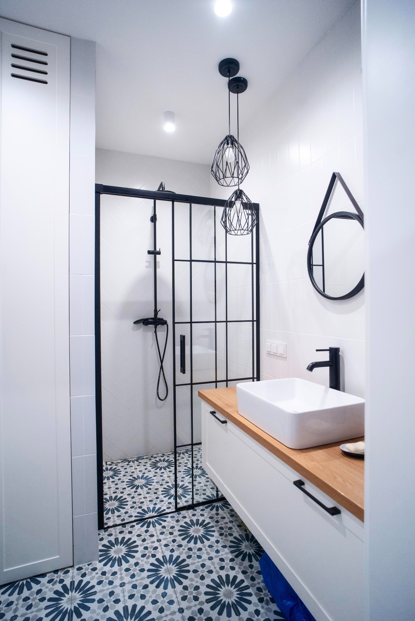 Nowoczesna łazienka z kabiną prysznicową typu walk-in z czarnymi ramami, umywalką nablatową na drewnianym blacie, geometrycznymi lampami wiszącymi i wzorzystą podłogą w odcieniach błękitu.