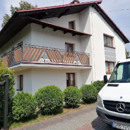 Dwukondygnacyjny dom jednorodzinny z białymi ścianami, drewnianymi oknami i balkonem z ozdobną balustradą, widok z poziomu kostki brukowej, z częściowo widocznym białym samochodem dostawczym.