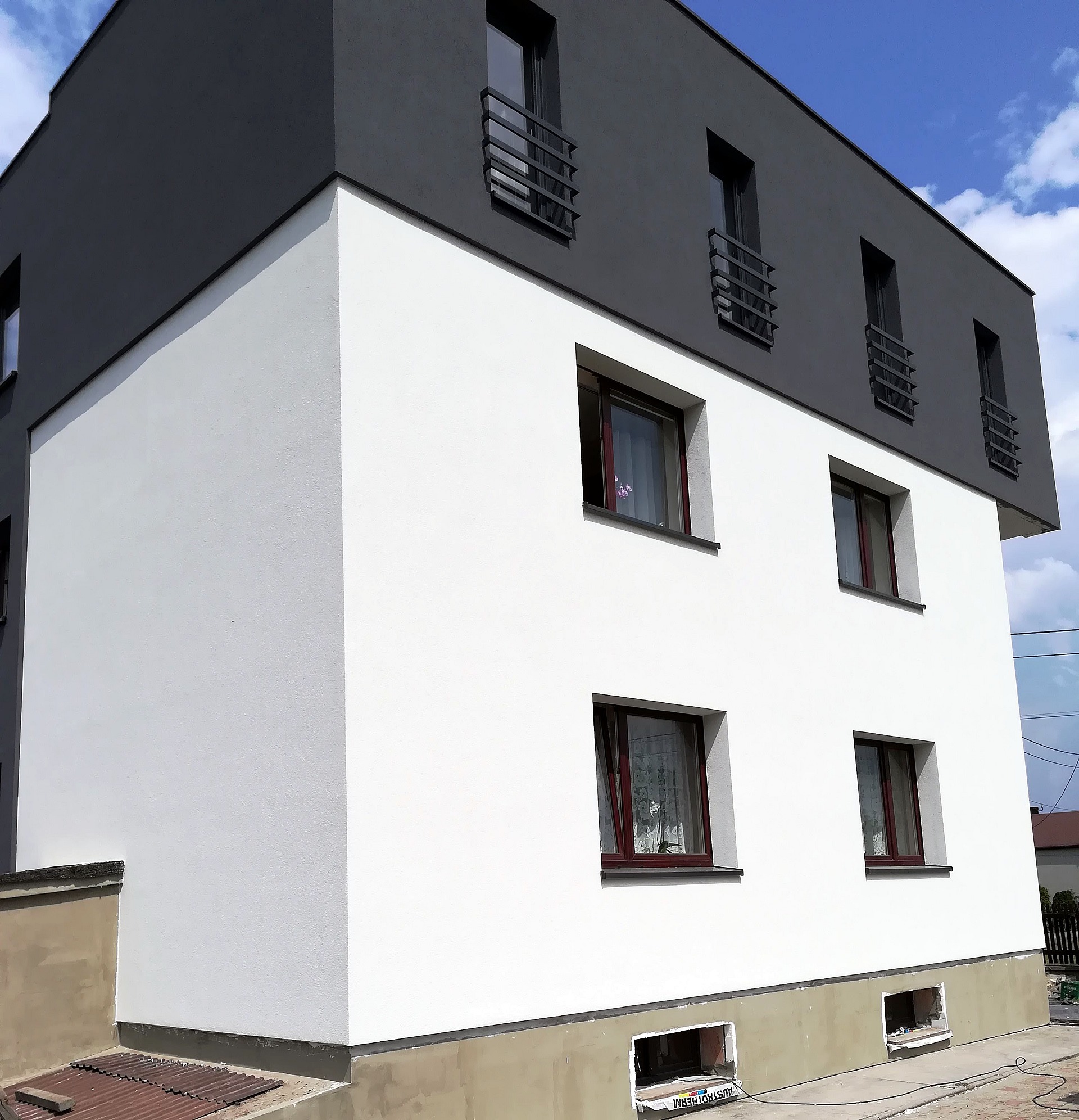 Dwukolorowa elewacja budynku po remoncie: biała ściana parteru i szara ściana piętra z oknami z balustradami, widoczne prace wykończeniowe przy fundamentach.