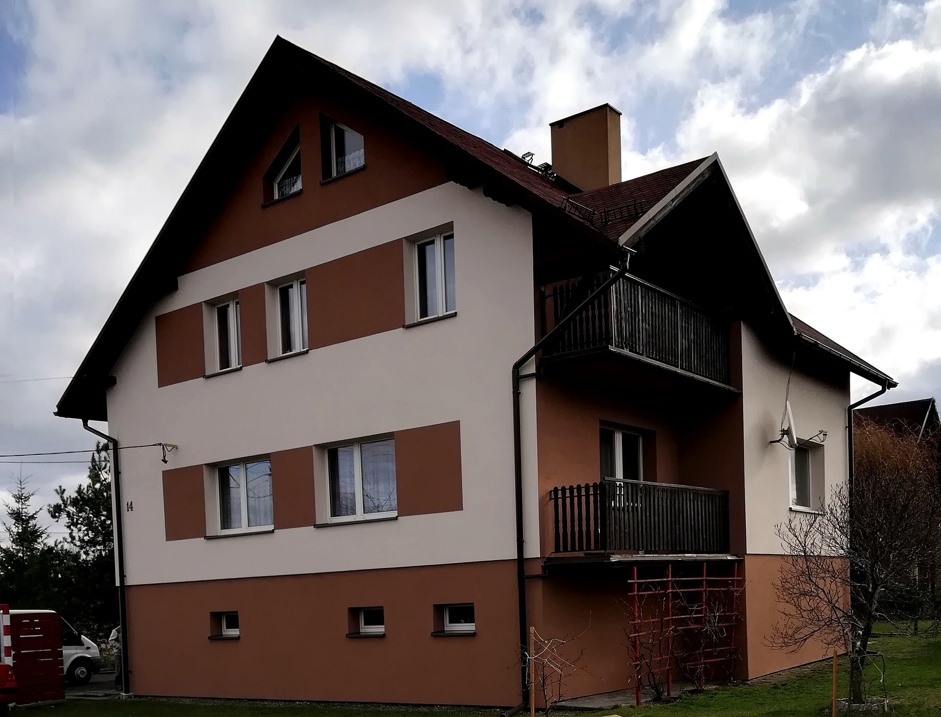 Dwukondygnacyjny dom z balkonem, wykończony w odcieniach brązu i beżu, z numerem 14 na ścianie, widoczny fragment ogrodu i samochodu dostawczego.