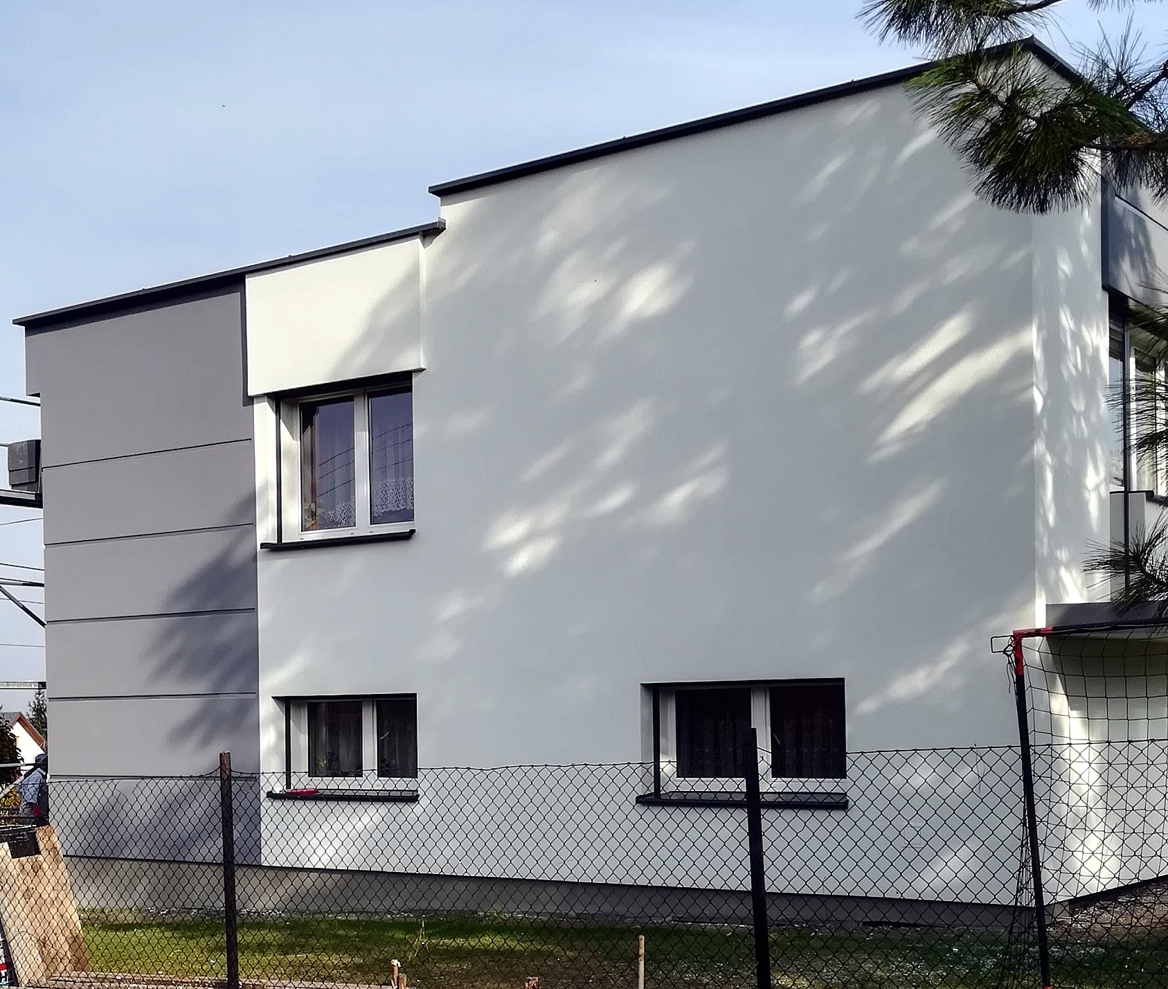 Dwukondygnacyjny budynek z fragmentem szarej elewacji panelowej i białą, gładką elewacją, widoczny przez siatkę ogrodzeniową i gałąź drzewa rzucającą cień.