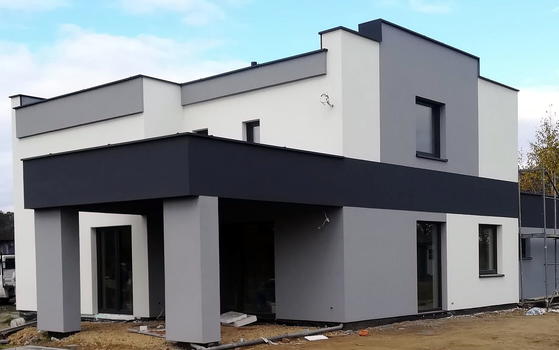 Nowoczesny dom jednorodzinny z elewacją w odcieniach szarości i bieli, widoczne rusztowania z boku budynku.