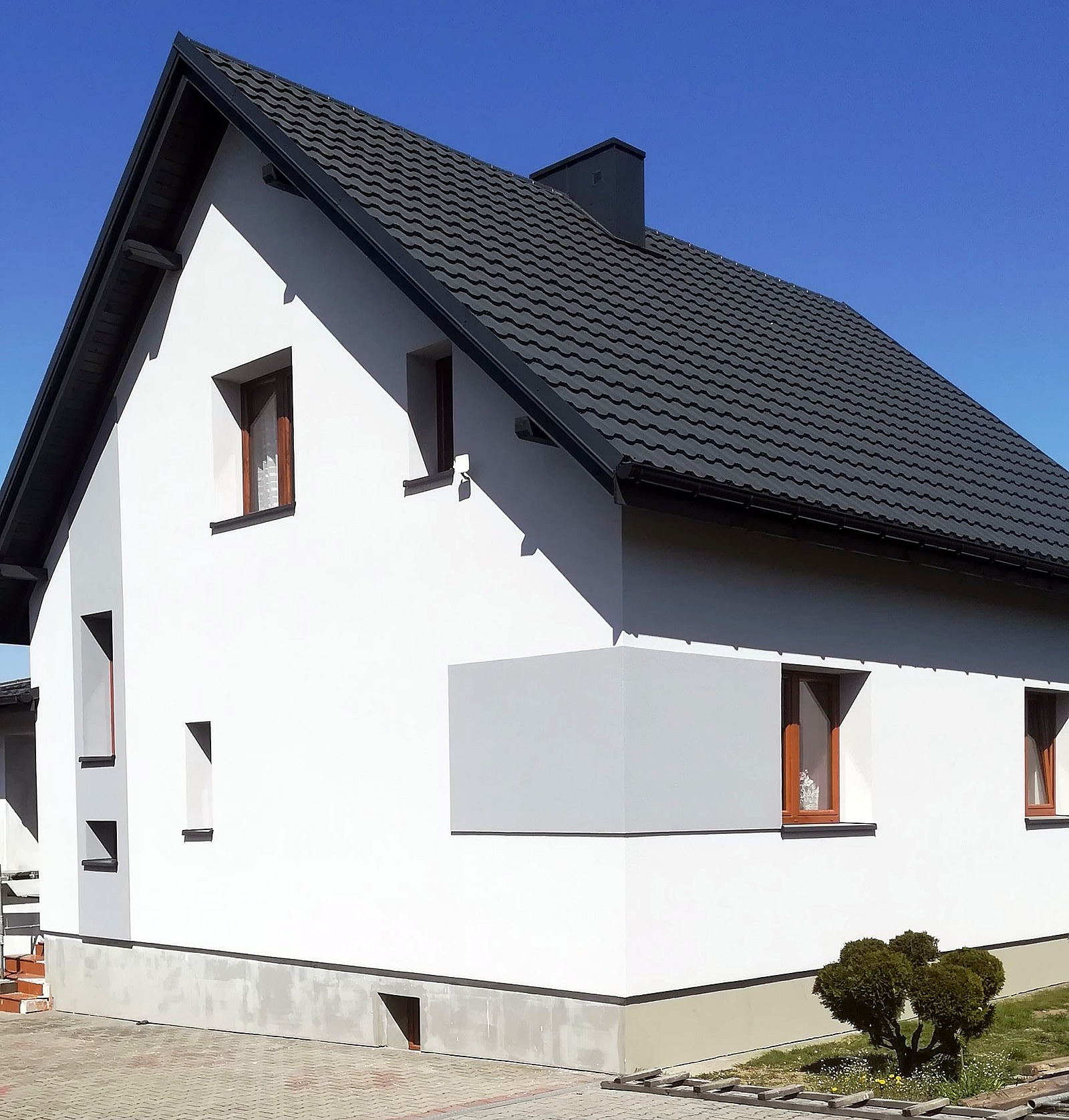 Nowoczesny dom jednorodzinny z białą elewacją i grafitowym dachem, widoczne okna z brązowymi ramami i fragment posesji z kostki brukowej.