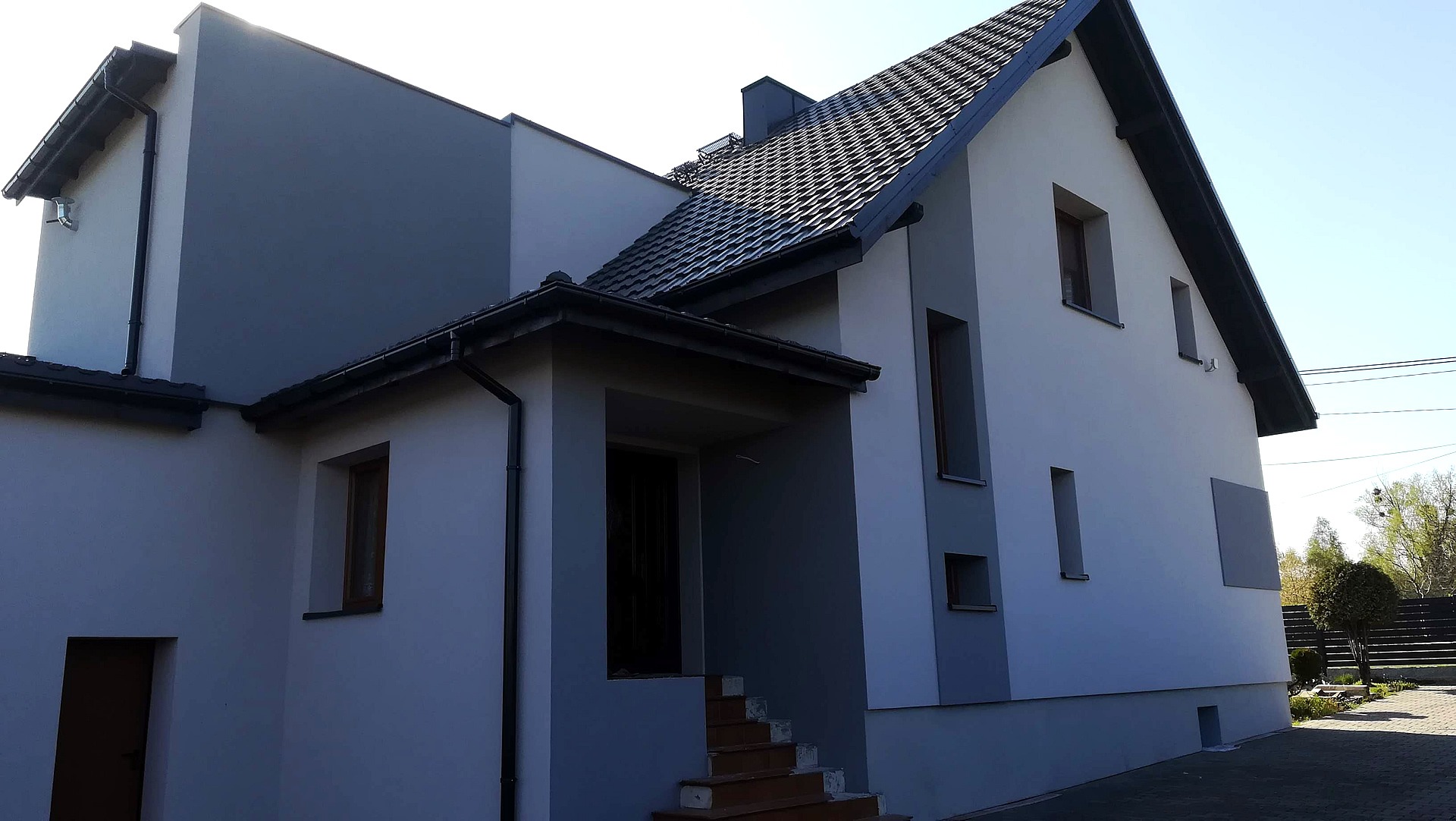 Dwukondygnacyjny dom z szarą i białą elewacją, ciemną dachówką i schodami wejściowymi, sfotografowany w słoneczny dzień.