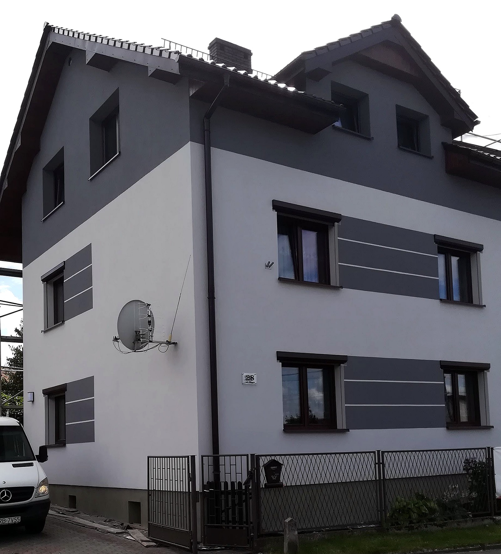 Dwukondygnacyjny dom z szaro-białą elewacją, z widoczną anteną satelitarną, brązowymi oknami i czarnym ogrodzeniem, sfotografowany z poziomu ulicy.
