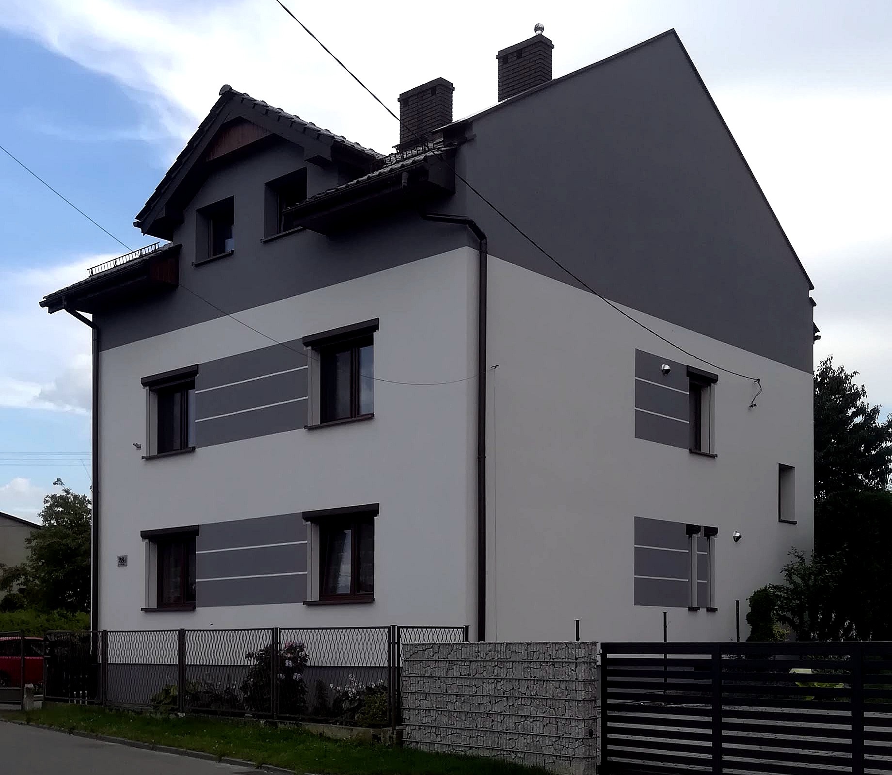 Dwukondygnacyjny dom z szarą górną częścią elewacji i białym dołem, z poziomymi szarymi pasami pod oknami, widoczny z ulicy za ogrodzeniem.