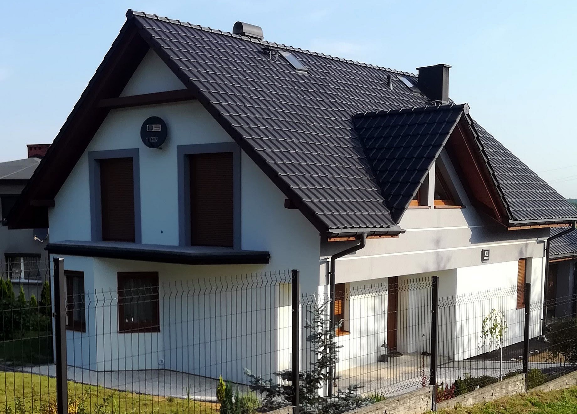 Nowoczesny dom jednorodzinny z szarą dachówką i jasną elewacją, widok z zewnątrz przez metalowe ogrodzenie.