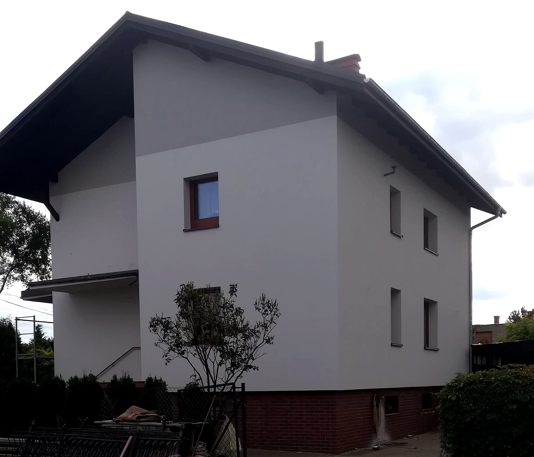 Dwukondygnacyjny dom z elewacją w odcieniach szarości i białym tynkiem, z widoczną ceglaną podmurówką i małym drzewkiem na pierwszym planie.