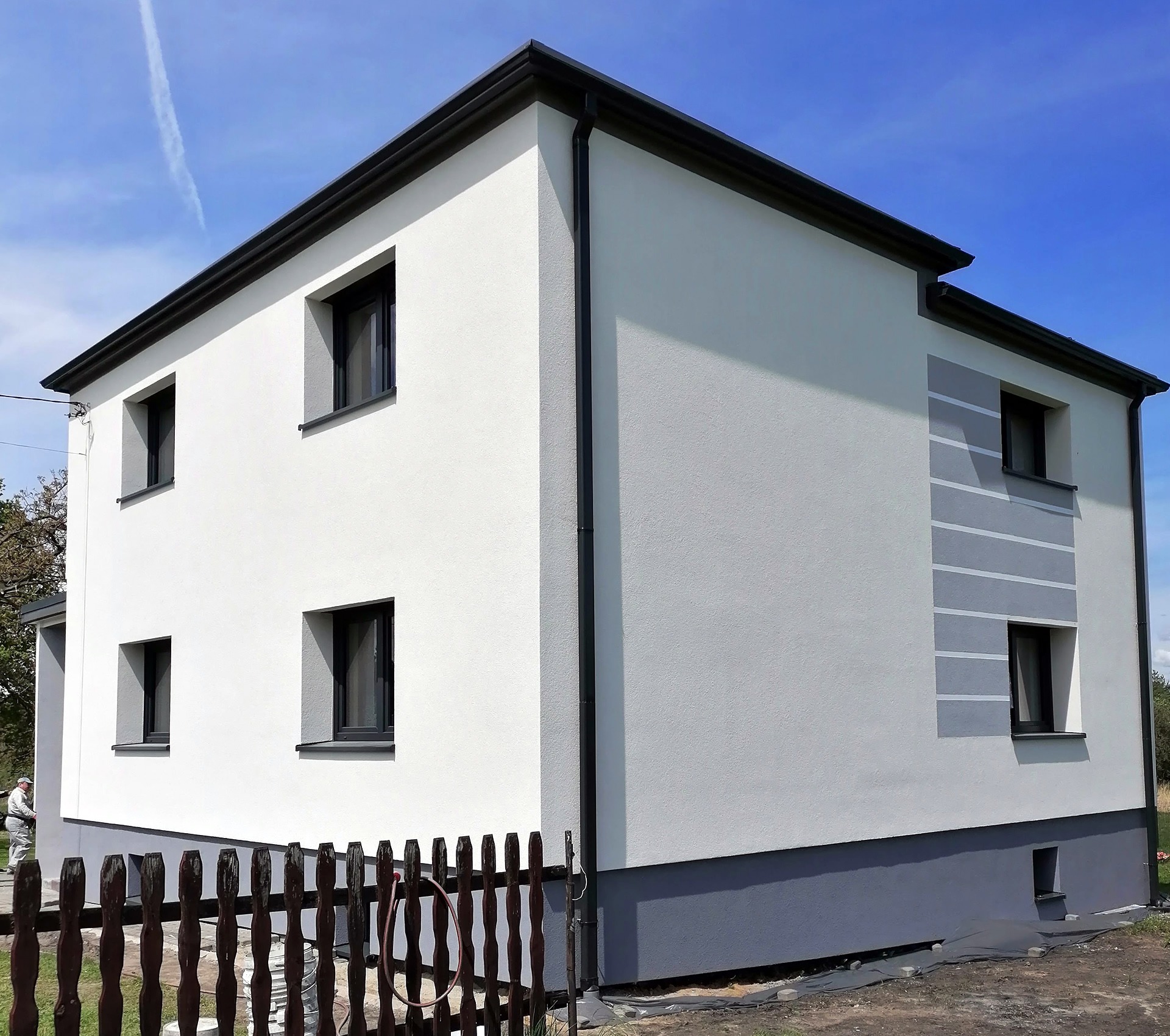Dwukondygnacyjny dom po remoncie elewacji, z białymi ścianami i grafitowymi detalami, widoczny fragment ogrodzenia i pracownik w tle.