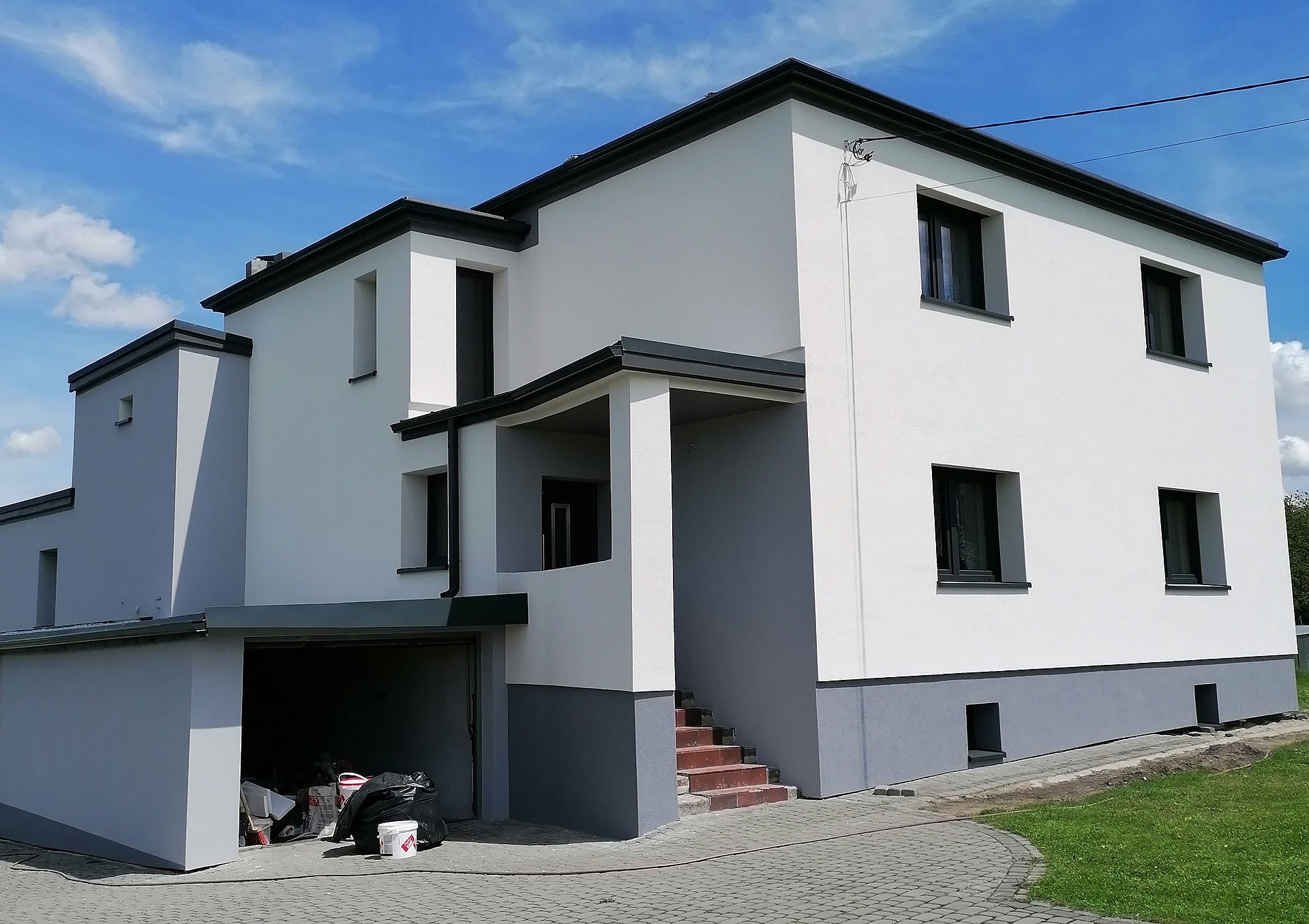 Nowoczesny dom jednorodzinny w Jejkowicach z białą elewacją i grafitowymi akcentami, widok z poziomu chodnika z kostki brukowej, otwarty garaż z widocznymi narzędziami i materiałami budowlanymi.