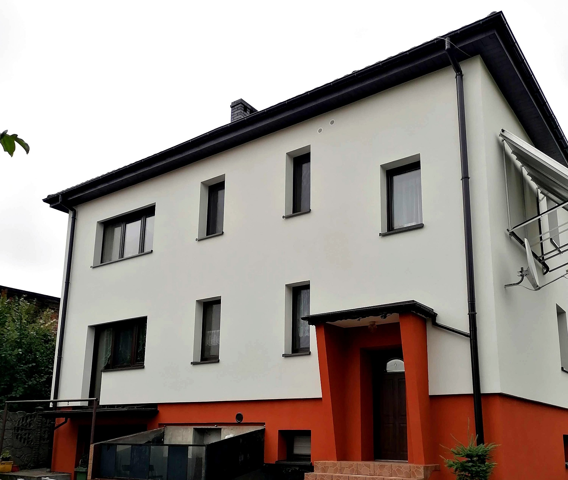 Dwupiętrowy dom po remoncie elewacji z białymi ścianami, pomarańczowym cokołem i obramowaniem wejścia, czarnymi oknami i rynnami.