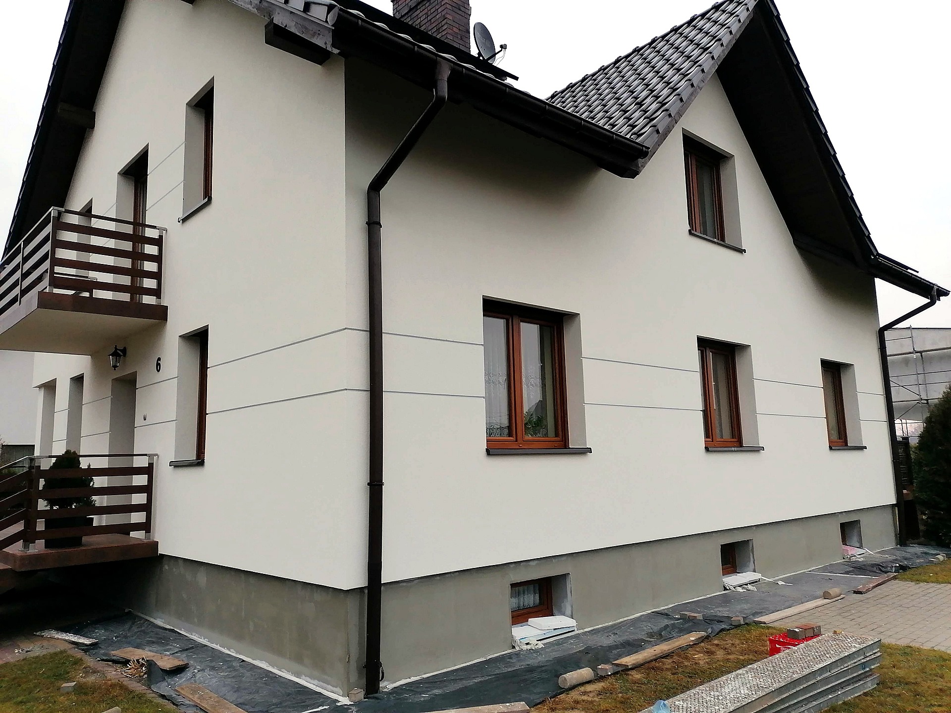 Elewacja domu jednorodzinnego po tynkowaniu i malowaniu, z widocznym fragmentem ocieplenia przy gruncie i przygotowanymi materiałami budowlanymi.