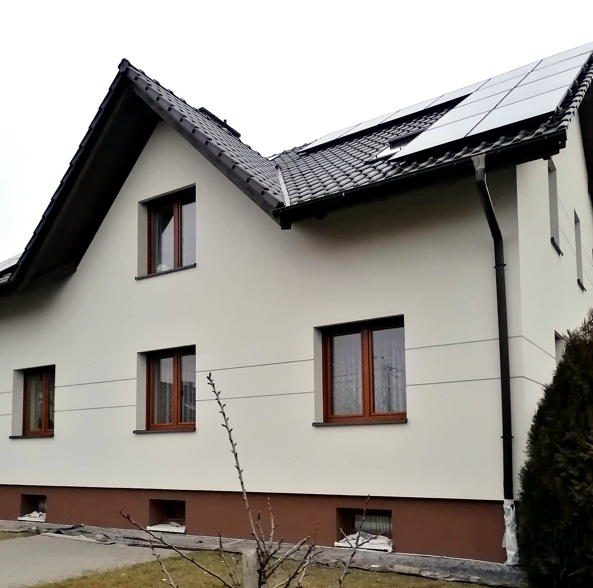 Dom jednorodzinny z jasną elewacją, brązowymi oknami i panelami słonecznymi na dachu, widok z zewnątrz w pochmurny dzień.