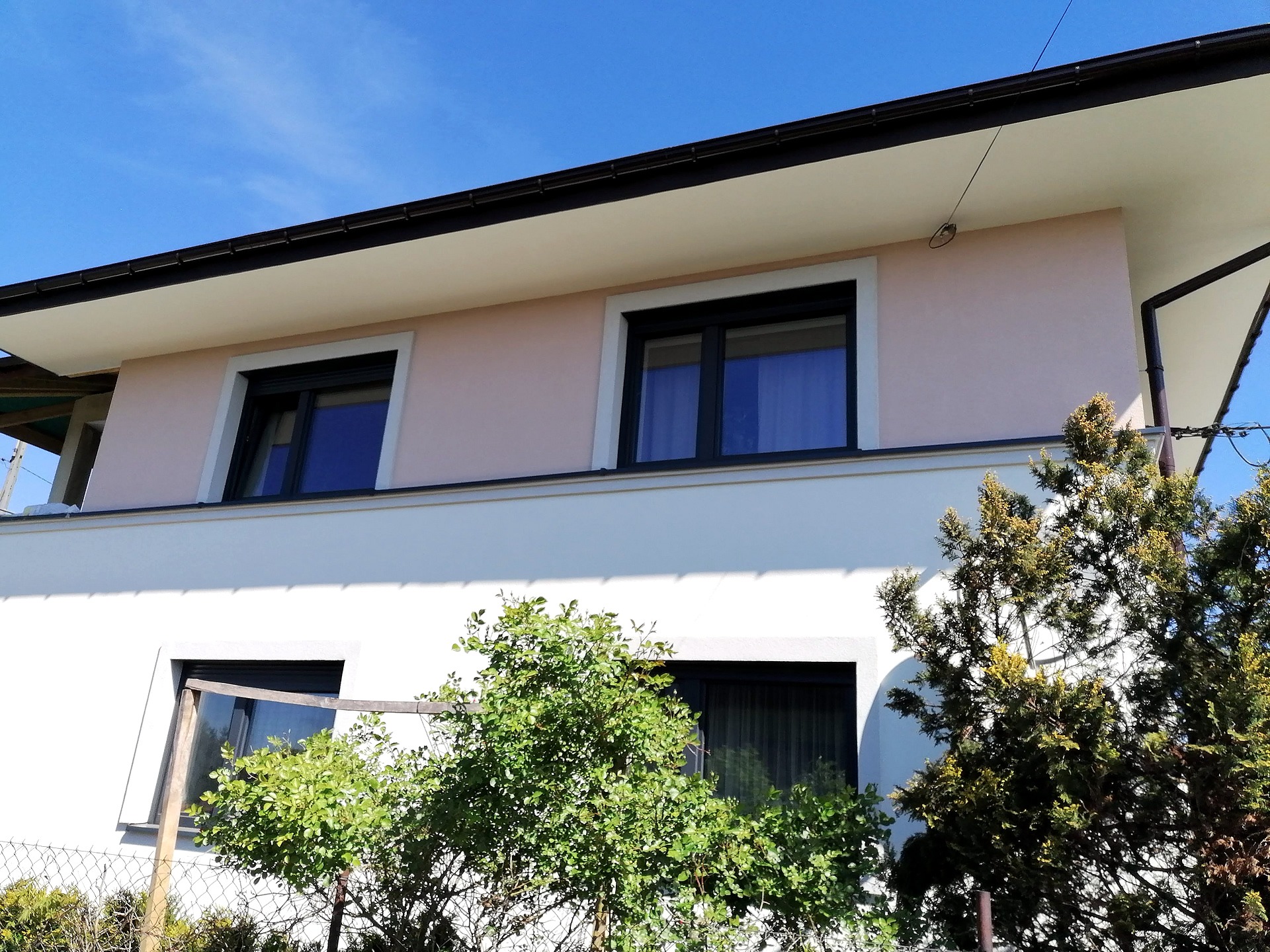 Dwukondygnacyjny dom z elewacją w odcieniach bieli i pudrowego różu, z czarnymi ramami okiennymi, widoczny spod kątem, częściowo zasłonięty zielenią.