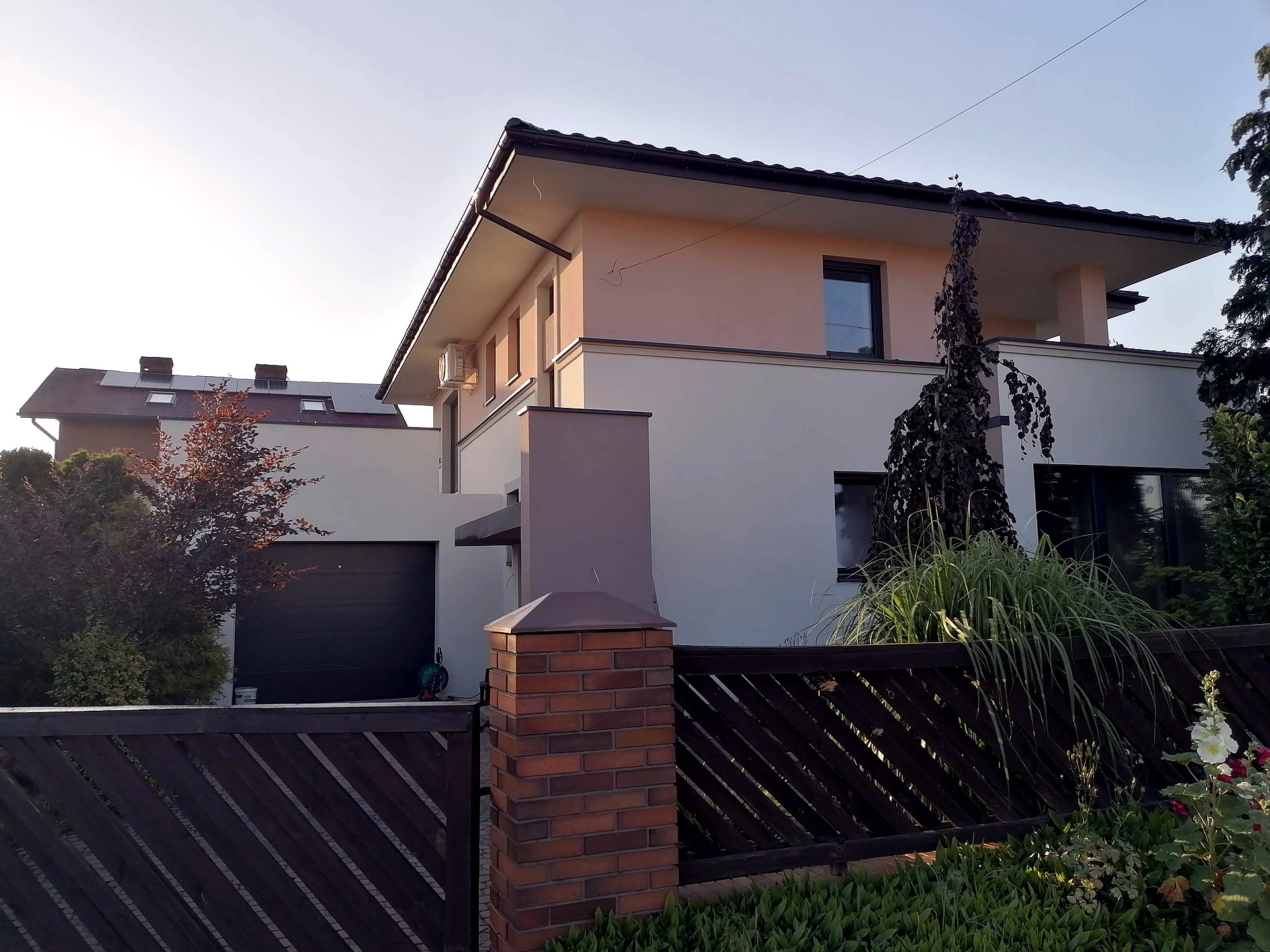 Dwukondygnacyjny dom jednorodzinny z płaskim dachem, elewacja w kolorze beżowo-białym, widok zza drewnianego ogrodzenia z ceglanym słupkiem, widoczny klimatyzator na ścianie.