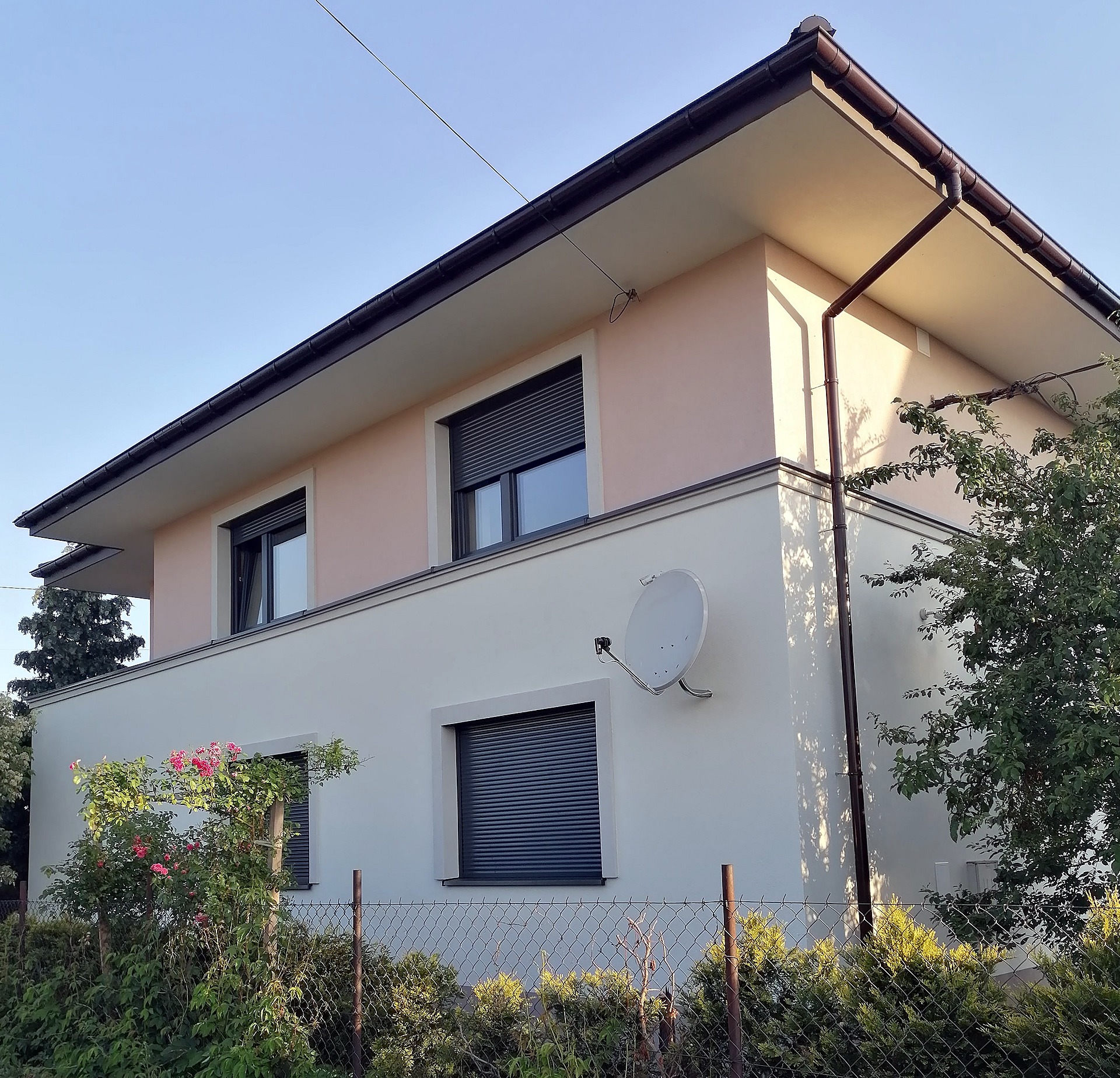 Dwukondygnacyjny dom z elewacją w odcieniach beżu i bieli, z roletami zewnętrznymi w oknach i anteną satelitarną na ścianie, otoczony zielenią.