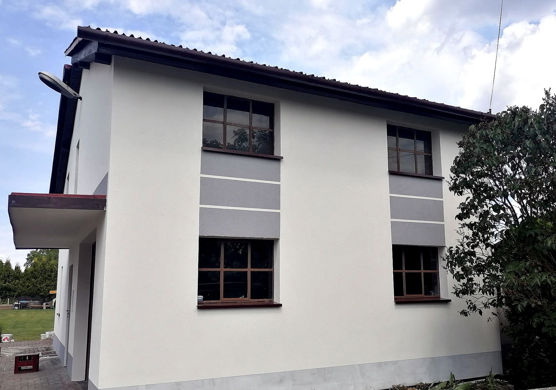 Dwukondygnacyjny budynek z jasną elewacją, szarymi pasami pod oknami i brązowymi ramami okiennymi, widoczne wejście z zadaszeniem i fragment ogrodu.