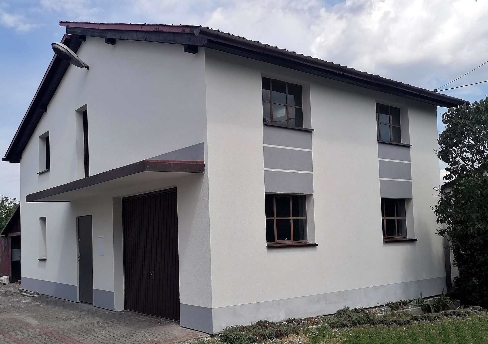 Dwukondygnacyjny dom z garażem, świeżo po remoncie elewacji, z szarymi akcentami wokół okien i u podstawy, oświetlony lampą zewnętrzną.