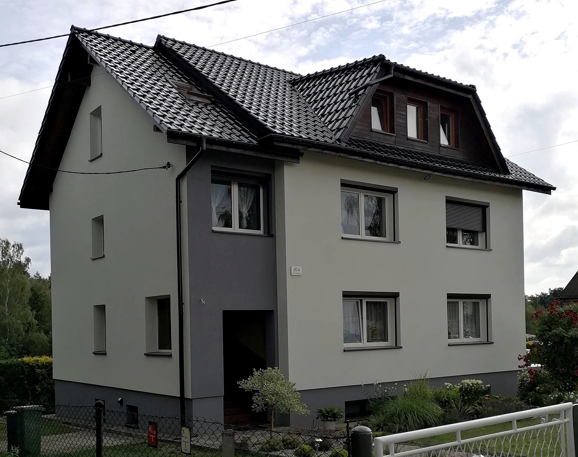 Dwukondygnacyjny dom z szarą elewacją i ciemną dachówką, widoczny z perspektywy ogrodu, z numerem domu 104.
