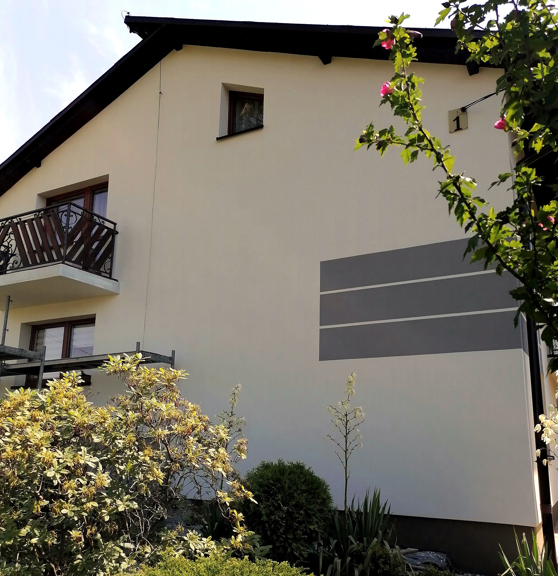 Elewacja domu jednorodzinnego po remoncie z dekoracyjnymi, szarymi pasami, widoczny balkon z ozdobną balustradą, rusztowanie i fragment bujnej roślinności.