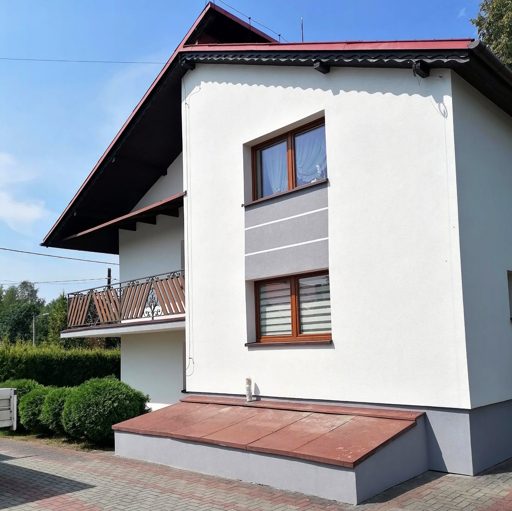 Biała elewacja domu z brązowymi oknami, szarym pasem dekoracyjnym i czerwoną dachówką, widok z zewnątrz, z balkonem i zielonymi krzewami w tle.