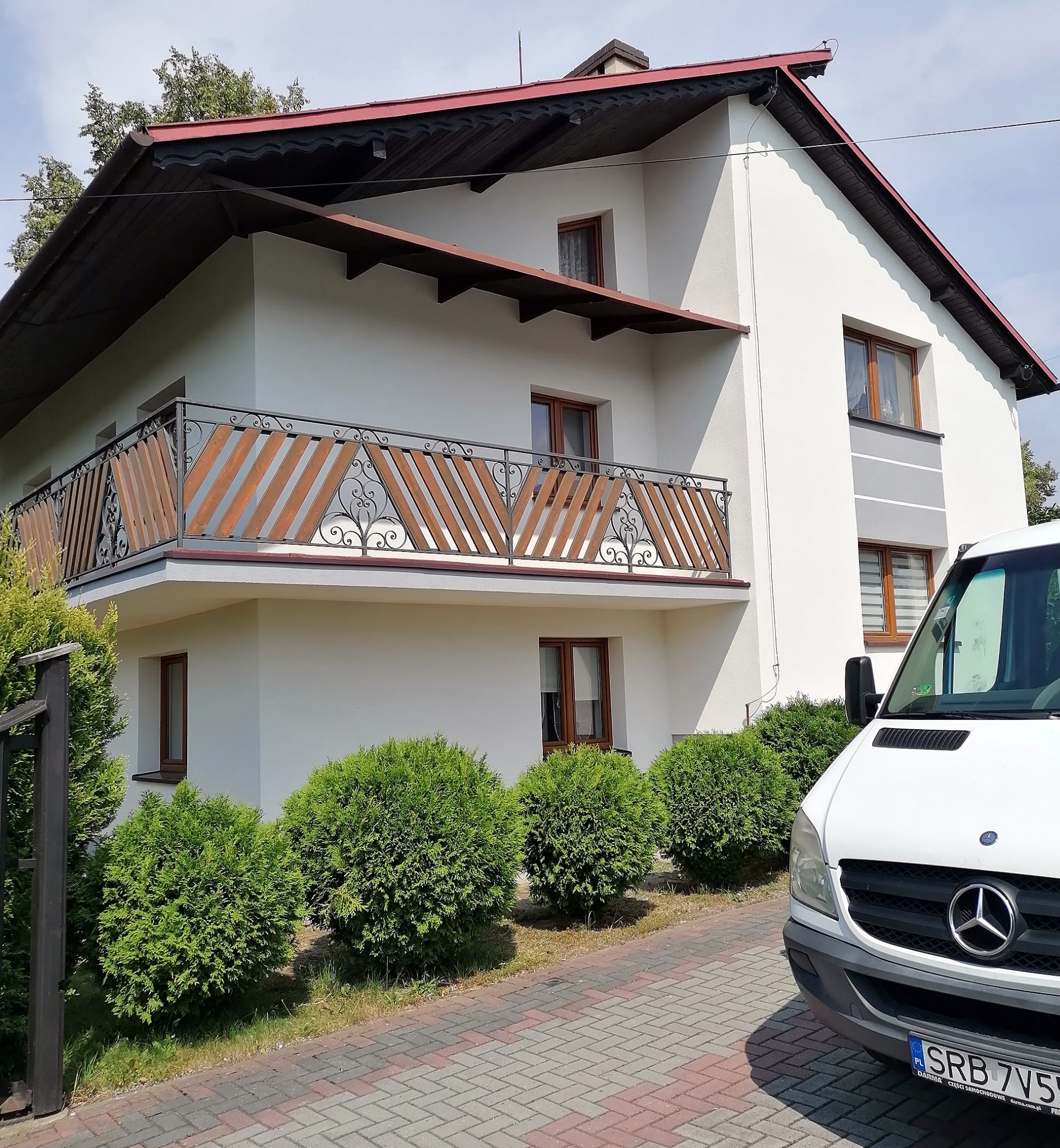 Dwukondygnacyjny dom jednorodzinny z białymi ścianami, drewnianymi oknami i balkonem z ozdobną balustradą, widok z poziomu kostki brukowej, z częściowo widocznym białym samochodem dostawczym.