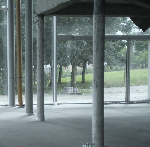Wnętrze budynku w stanie surowym zamkniętym, z widoczną szarą, gładką posadzką, okrągłymi kolumnami częściowo owiniętymi taśmą i dużymi oknami z widokiem na zieleń.
