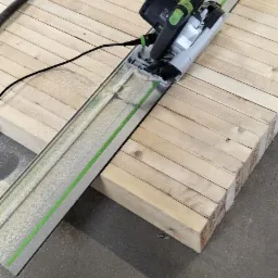 Piła zagłębiarka Festool na szynie tnąca drewniane deski, widoczne ślady cięcia i pył.