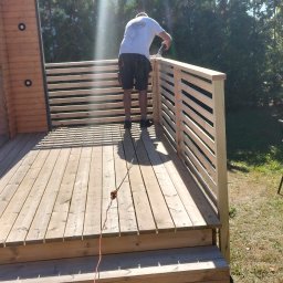 D-Wood - Montaż drewnianego tarasu przy domu z drewnianą elewacją, widoczny pracownik podczas instalacji balustrady z poziomych desek, w tle stół z krzesłami na trawniku.