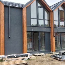 D-Wood - Wznoszenie nowoczesnego domu z dużymi oknami, pracownicy na drabinie i przy samochodzie dostawczym, widoczne materiały budowlane.