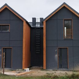 D-Wood - Szereg identycznych, nowoczesnych domów z ciemną elewacją i drewnianymi elementami wykończeniowymi, połączonych metalową drabinką na dachu, widoczne ślady budowy.