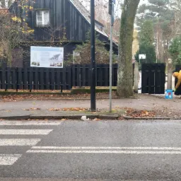 Malowanie czarnej, drewnianej bramy wejściowej na posesję z czarnym domem, widoczne wiadro z farbą i dwie osoby obserwujące.