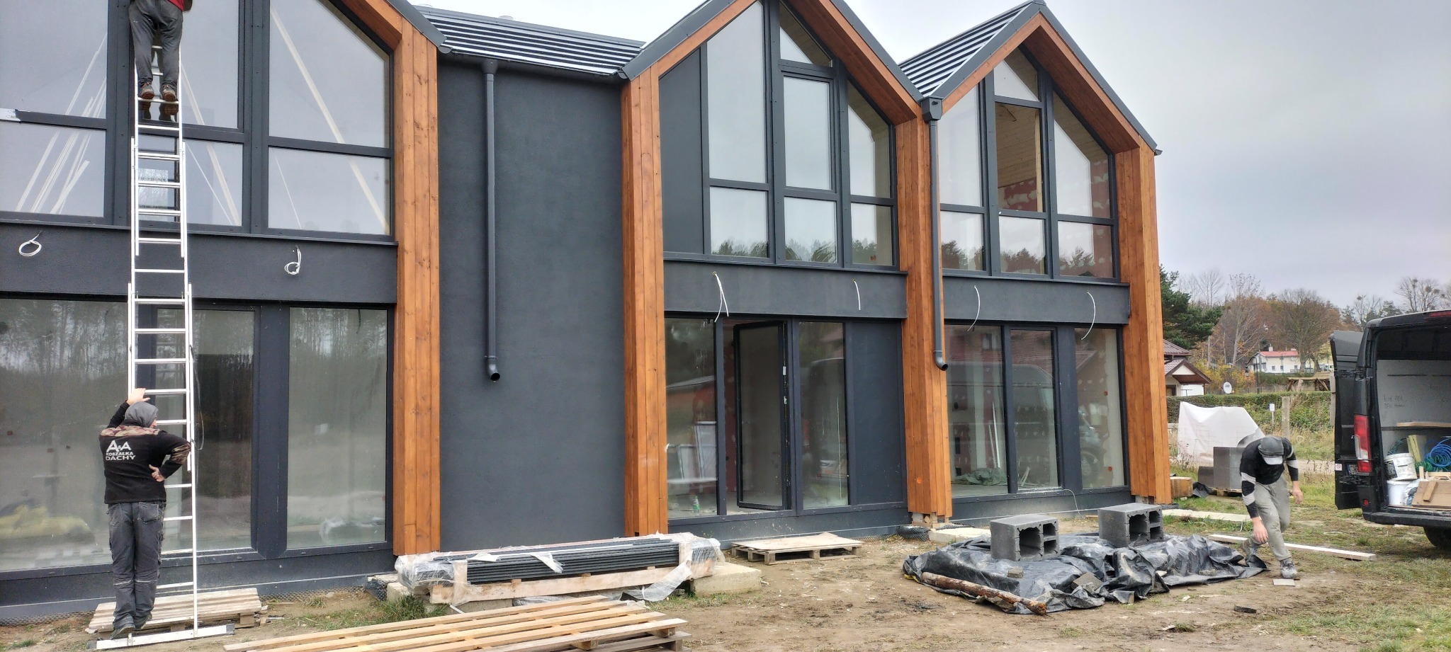 Wznoszenie nowoczesnego domu z dużymi oknami, pracownicy na drabinie i przy samochodzie dostawczym, widoczne materiały budowlane.