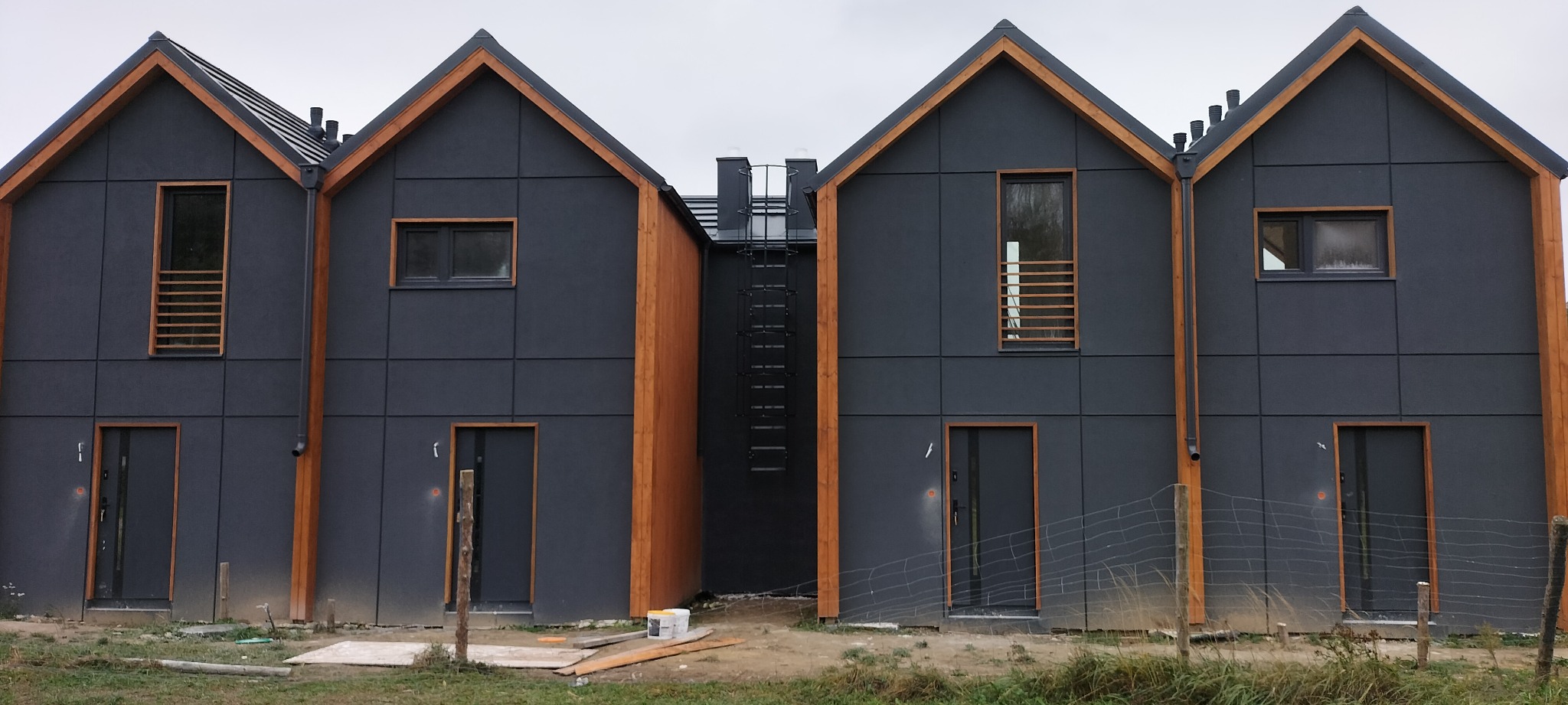 Szereg identycznych, nowoczesnych domów z ciemną elewacją i drewnianymi elementami wykończeniowymi, połączonych metalową drabinką na dachu, widoczne ślady budowy.