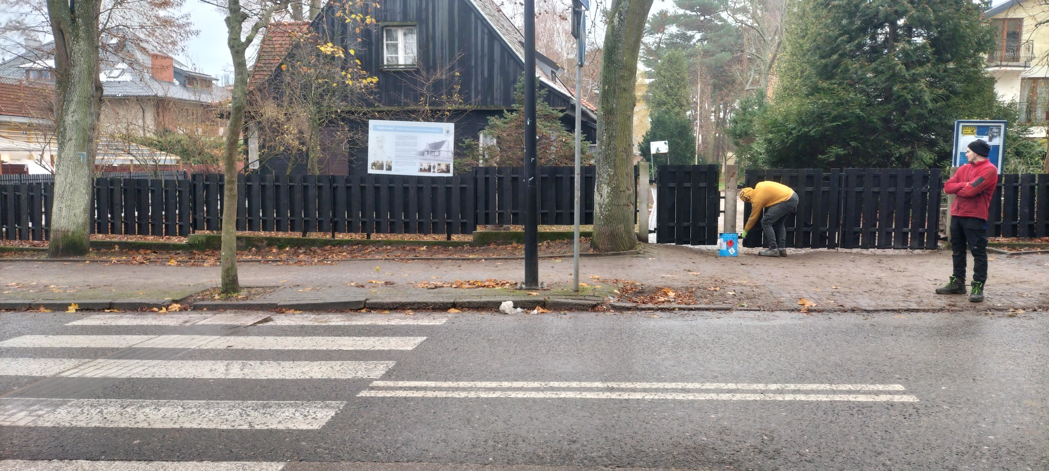 Malowanie czarnej, drewnianej bramy wejściowej na posesję z czarnym domem, widoczne wiadro z farbą i dwie osoby obserwujące.