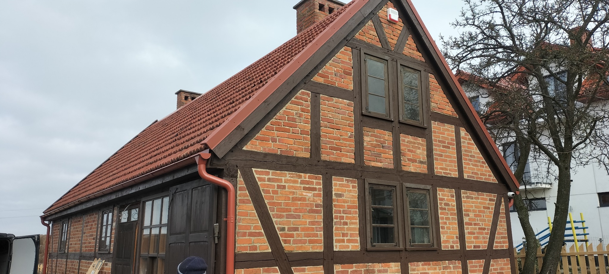 Elewacja budynku z czerwonej cegły z drewnianą konstrukcją szkieletową, widoczne okna i dach z czerwonej dachówki, na zewnątrz rynna w kolorze dachu.