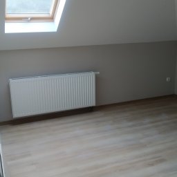 Dmytro Protsenko - Poddasze ze skośnym sufitem, drewnopodobną podłogą, białym grzejnikiem i oknem dachowym, ściany pomalowane na dwa odcienie szarości.